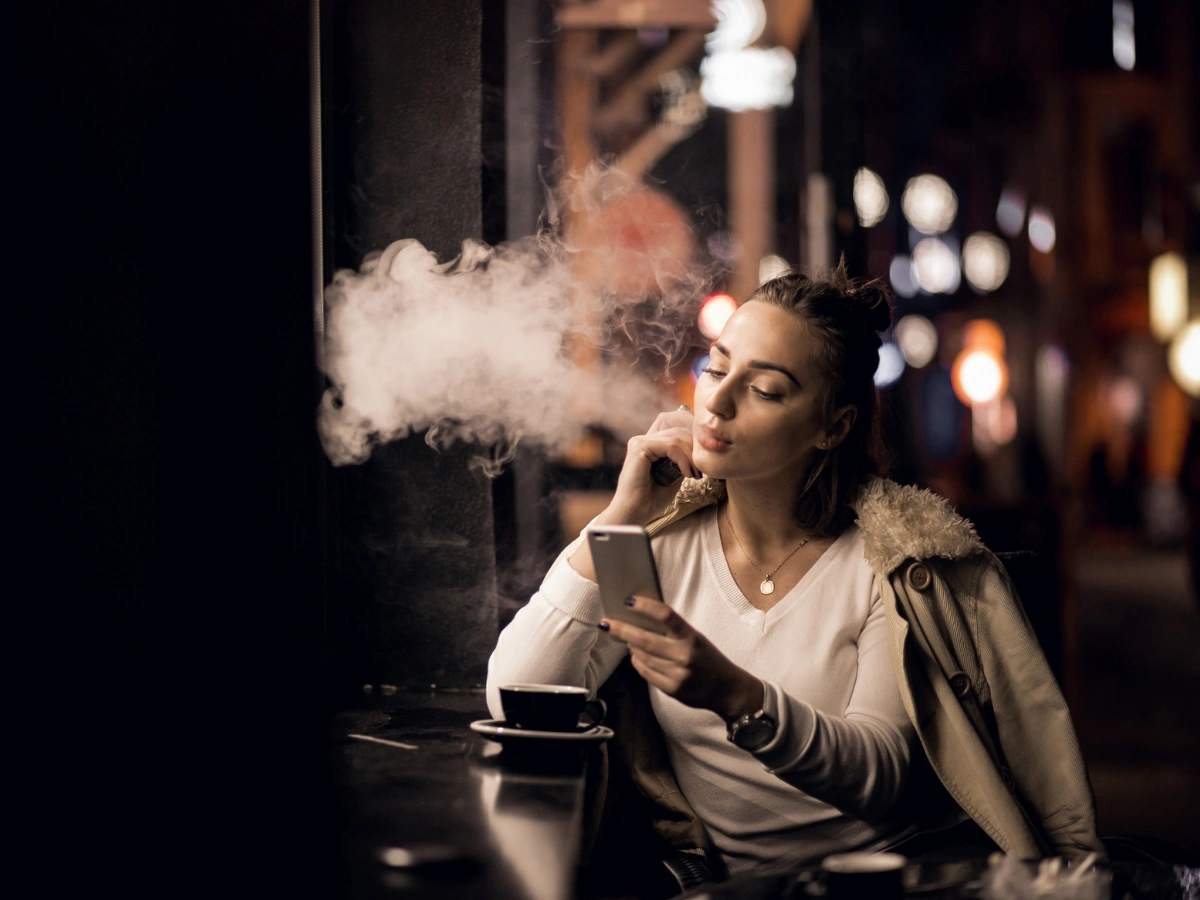 Woman using vape mod while checking phone in a café – Vape Mods for Beginners.